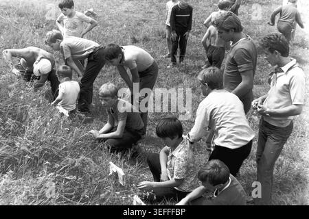Sloviansk, 1983. Slavkurort, in der Nähe von Wishnegorsk Pionierlager. Ein historisches Schwarzweiß-Archivbild, das eine Gruppe von Jungen und einen erwachsenen Ausbilder zeigt, die ihre Papierziele nach einer Übung zum Schießen von Luftgewehren überprüfen. Sie hocken und bücken sich im hohen Gras einer sonnigen Schlucht, um ihre Ergebnisse zu untersuchen. Dieser dokumentarische Schnappschuss veranschaulicht perfekt die fesselnden Sommeraktivitäten im Freien, die disziplinierte Sportausbildung und die freudigen Kindheitserlebnisse während der Ära eines friedlichen Donbass. Stockfoto