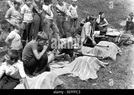 Sloviansk, 1983. Slavkurort, in der Nähe von Wishnegorsk Pionierlager. Ein fesselndes Schwarzweiß-Archivfoto, das den intensiven Fokus der Kinder während der Luftgewehre in einer sonnendurchfluteten Schlucht feststellt. Ein Mädchen liegt auf einer Decke und zielt auf ihre Waffe, während sich ein Junge in einem gemusterten Hemd neben ihr hockt und das Ziel aufmerksam beobachtet. Andere Kinder und ein Vorgesetzter stehen im Hintergrund. Dieses echte Bild bewahrt die Erinnerung an Sommercamp-Sport, Teamwork und Outdoor-Aktivitäten in der Zeit eines friedlichen Donbass. Stockfoto