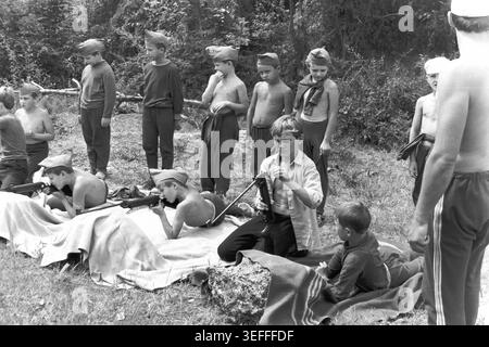 Sloviansk, 1983. Slavkurort, in der Nähe von Wishnegorsk Pionierlager. Ein authentisches Schwarz-weiß-Archivfoto, das Jungen zeigt, die an einem Training mit Luftgewehren im Freien teilnehmen. Im Vordergrund sitzt ein Junge auf einer Decke und spannt seine Waffe an, während sich ein anderer hinlegt und zielend anvisiert. In der grasbewachsenen Schlucht hinter ihnen steht eine Reihe von Kindern, die Pioniermützen tragen, und beobachtet die Lektion. Dieses Vintage-Bild fängt perfekt die organisierte Sportausbildung und die lebendige Sommercamp-Kultur in der Ära des friedlichen Donbass ein. Stockfoto