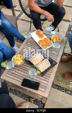 Ein hoher Winkel von nicht erkennbaren Leuten, die während eines auf einem Holztisch erfrischende Cocktails wie Margaritas, Mini-Thunfisch-Sandwiches, Slider und Chips genießen Stockfoto