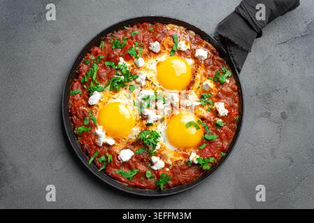 Köstliche Shakshuka: Spiegeleier mit Tomaten, Fetakäse und Petersilie in einer gusseisernen Pfanne. Frisches Shakshuka – mediterranes Helthy-Frühstück auf Steinplatte Stockfoto