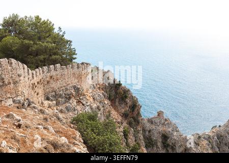 Die historische Steinfestungsmauer steht auf einer felsigen Klippe, umgeben von Grün, mit einem Panoramablick auf das ruhige Meer im Hintergrund, das die Natur zeigt Stockfoto