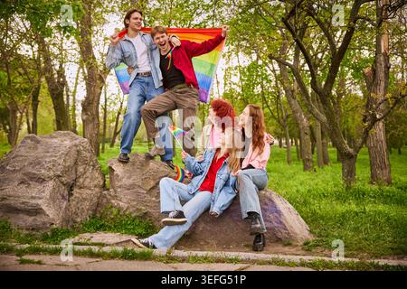 Junge Freunde feiern mit Regenbogenfahne auf Felsen im sonnigen Park. Stockfoto