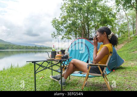 Schwarze Frau, die Fotos mit der Kamera macht, während sie am See in der Natur campt Stockfoto