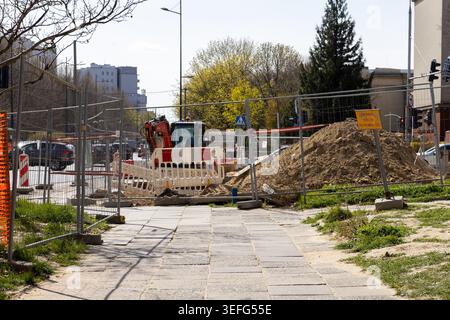 Ein kleiner Bagger steht auf einem Schotterhaufen hinter temporären Metallbarrieren auf einer städtischen Straßenbaustelle und veranschaulicht die Stadtentwicklung, die Versorgungsanlagen Stockfoto