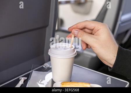 Eine Nahaufnahme einer Person, die einen Holzstock verwendet, um ein Getränk in einem weißen Tassen zum Mitnehmen auf einem Sitztablett des Flugzeugs zu rühren, was die Bequemlichkeit von tra unterstreicht Stockfoto