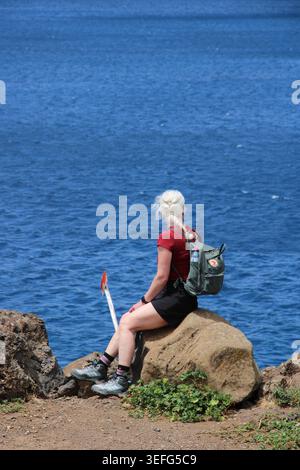 Frau mit weißem Haar sitzt auf felsigem Felsvorsprung. Sie blickt auf das weite blaue Meer, ruhig und nachdenklich. Rucksack und Wanderausrüstung bieten Outdoor-Abenteuer. Sonne Stockfoto