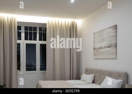 Minimalistisches Schlafzimmer mit einem weichen Bett, abstrakten Wandbildern und drapierten Fenstern, die Komfort und Raffinesse für die Innenarchitektur vermitteln Stockfoto