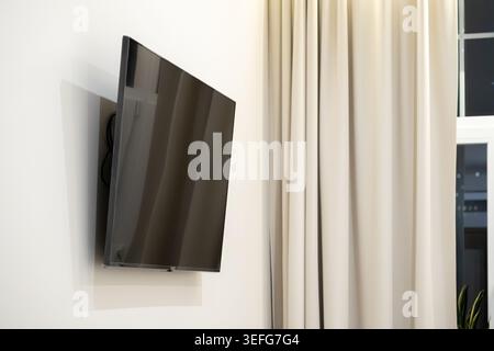 Schwarzer LED-Fernseher, der in einem Winkel neben eleganten Vorhängen in einem hellen Apartment wandmontiert ist, veranschaulicht den zeitgenössischen Medienstil, Smart Hom Stockfoto