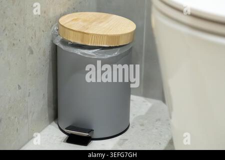 Kleiner Abfallbehälter im Badezimmer mit schlanker matter Oberfläche und Deckel mit Holzakzenten, der auf Steinfliesen platziert wird, um organisierte Hygiene, nachhaltiges Wohnen, Stockfoto