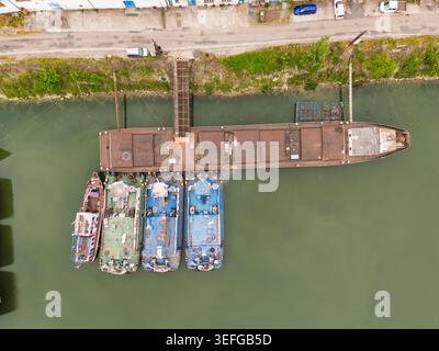 Aus der Vogelperspektive von Industriekähnen und Booten, die an einem Flussufer mit einer Gangway und grünem Wasser in Bratislava, Region Bratislava, Slowakei, vor Anker gestellt sind. Stockfoto