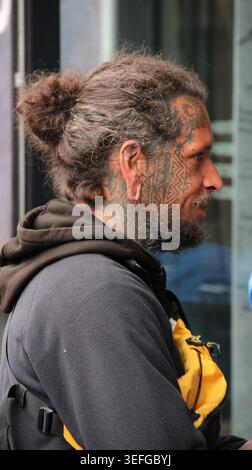 Mann mit Tattoos und langen Haaren in Brötchen. Er trägt einen dunklen Hoodie und eine gelbe Tasche. Sein Gesicht zeigt Ruhe, vielleicht Müdigkeit. Straßenszene hinter ihm, verschwimmt Stockfoto