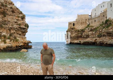 Entspannter Mann, der die malerische mediterrane Bucht mit felsigem Strand und Altstadt an einem bewölkten Sommertag erkundet. Stockfoto