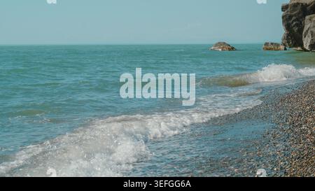 Schäumende weiße Wellen krachen auf einen Kieselstrand unter einem klaren blauen Himmel Stockfoto