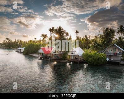 Blick aus der Vogelperspektive auf tropische Bungalows entlang der türkisfarbenen Küste, umgeben von üppigen Palmen unter einem glühenden Sonnenuntergangshimmel Moorea-Maiao, Windward Island Stockfoto