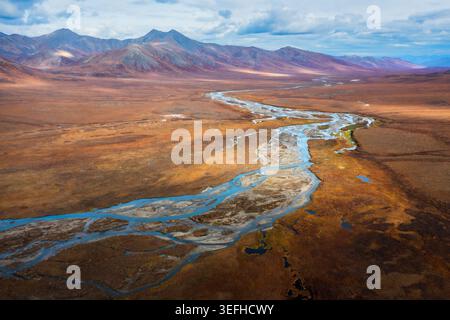 Aus der Vogelperspektive eines geflochtenen Flusses, der sich durch das weite herbstliche Tundra-Tal in der Nähe des Dalton Highway mit zerklüfteten Bergen unter einem bewölkten Himmel in Coldfoot schlängelt. Stockfoto