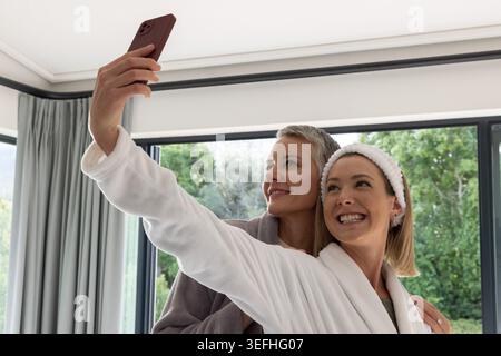 Seniorenmutter und Erwachsene Tochter machen zu Hause Selfie in der Nähe von Glastüren, tragen Bademäntel mit Telefon Stockfoto