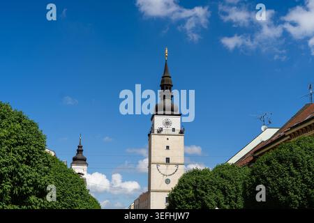 Blick auf den historischen Stadtturm mit seinem unverwechselbaren dunklen Turm und der goldenen Statue, die von üppigen grünen Bäumen unter einem blauen Himmel in Trnava, Region Trnava, eingerahmt wird Stockfoto