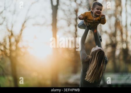 Mutter hebt den Sohn des Kleinkindes während der goldenen Stunde in einem Park in die Luft, ein freudiger Familienmoment Stockfoto