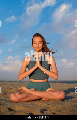 Eine Frau sitzt in padmasana auf dem Sand am Strand und spielt Namaste, während die Sonne im Hintergrund untergeht, was eine ruhige Atmosphäre schafft. Stockfoto