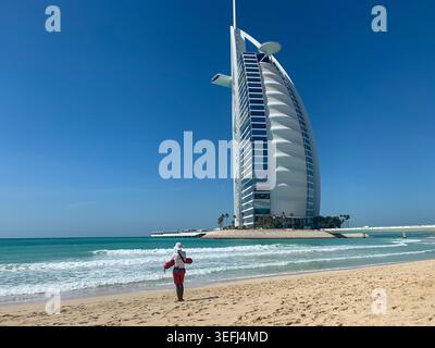 Blick auf das Burj Al Arab Hotel mit einem Rettungsschwimmer in roten Shorts am Sandstrand am türkisfarbenen Meer unter klarem blauen Himmel in Dubai, Dubai, Uni Stockfoto
