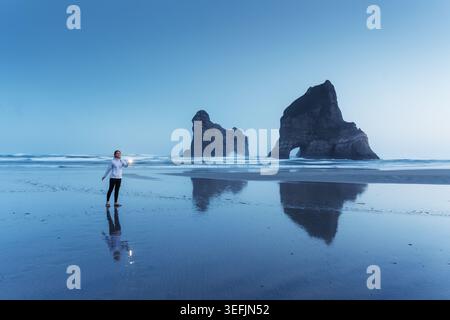 Eine ruhige, blaue Hour-Szene mit einer Frau, die am Wharariki Beach leuchtet, und die legendären Archway Islands reflektieren auf nassem Sand in Neuseeland Stockfoto