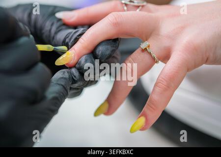 Nahaufnahme eines Nageltechnikers in schwarzen Handschuhen, der während einer professionellen Salonmaniküre einen gelben glänzenden lack auf einen langen Mandelnagel aufbürstet. Stockfoto