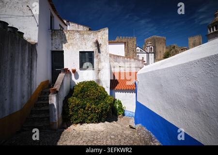 Traditionelle Kopfsteinpflasterstraße in Obidos mit weißen Häusern und mittelalterlichen Burgmauern, die hinter der historischen Stadt in Portugal aufragen. Stockfoto