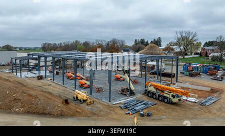 Bauarbeiter bauen mit Kränen und LKWs neue große Bauwerke. Der Standort ist mit Maschinen und Materialien beschäftigt. In der Nähe befinden sich Häuser und Bäume im Hintergrund. Stockfoto
