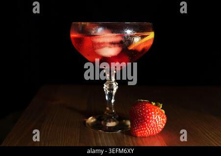 Erfrischender roter Cocktail in einem Glas mit Eis, Kirschen, Orangenschale und einer frischen Erdbeere auf dunkler Holzoberfläche, perfekt für Sommerabende und festliche Feierlichkeiten Stockfoto