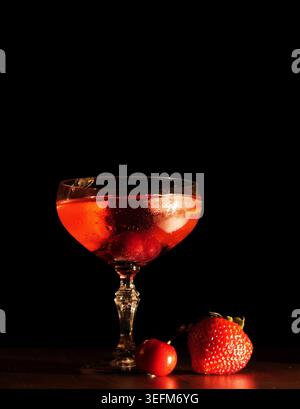 Cocktailglas gefüllt mit einem erfrischenden roten Getränk, garniert mit Kirschen und einer Orangenschale, ruht neben einer frischen Erdbeere und Kirsche im Dunkeln Stockfoto
