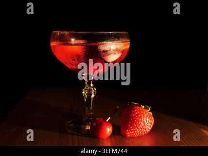 Ein Glas eisgekühlter roter Cocktail auf einer Holzoberfläche, garniert mit einer Reifen Kirsche und frischer Erdbeere, schafft ein lebendiges und erfrischendes Getränkeerlebnis Stockfoto
