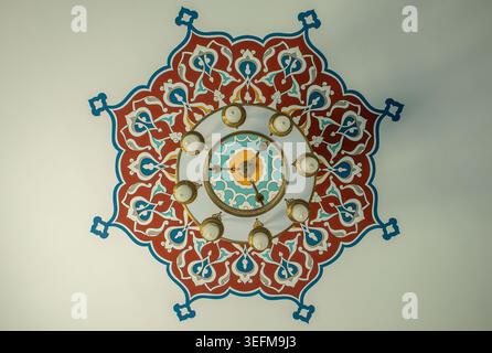 Ein symmetrischer niedriger Winkel auf eine farbenfrohe geometrische Deckendekoration mit einem Kronleuchter in einer modernen Moschee. Das Design hebt die Architektur hervor Stockfoto