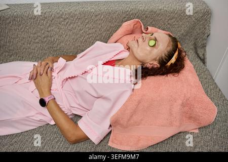 Eine reife Frau liegt auf einem Sofa mit einer Tonmaske und Gurkenscheiben über den Augen während einer Spa-Behandlung zu Hause Stockfoto