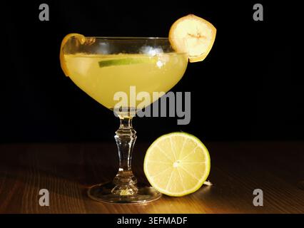 Blassgelbe Bananendaiquiri in einem Coupé-Glas mit Limettenscheibe und Bananengarnitur auf dunklem Holz vor schwarzem Hintergrund, tropischer Cocktail für den Sommer, S. Stockfoto