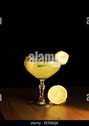 Cocktail mit raffinierten tropischen Aromen, einer Bananenscheibe und einer Limettenschale sowie einer frischen Limettenhälfte auf einem dunklen Holztisch Stockfoto