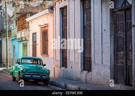 In Matanzas, Kuba, befindet sich ein blauer Oldtimer an einer historischen Straße. Die verwitterten Fassaden von Kolonialgebäuden und schmiedeeisernen Details schaffen einen Stockfoto