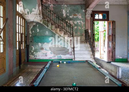 Ein abgenutzter Billardtisch steht in einem Kolonialraum in Kuba mit Marmortreppe und abblätternden grünen Wänden. Natürliches Licht strömt durch eine offene Tür Stockfoto