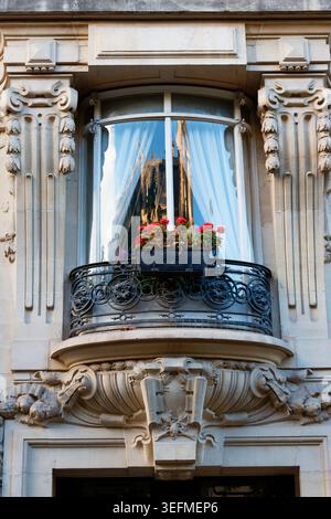 Der typisch Pariser Balkon mit Bugfenster ist mit leuchtenden roten Geranien in einem Fensterkasten geschmückt, eingerahmt von exquisiten Steinpilastern und einem Feinkostgeschäft Stockfoto