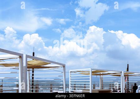 Eine moderne Bar-Dachterrasse in Penang, Malaysia, mit weißen Pergolen und beigefarbenen Segeln vor einem hellblauen Himmel mit weißen Wolken. Das Open-Air-Hotel blickt auf das Meer. Stockfoto