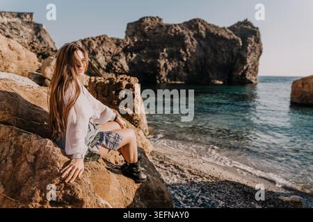 Frau Ocean Coast junge Frau in weißer Bluse, die auf zerklüfteten Felsen sitzt und das türkisfarbene Meer während der goldenen Stunde überblickt Stockfoto