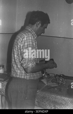 Sloviansk, 1980er Jahre, authentischer Werkstattbereich im Haus. Ein Vintage-Schwarz-weiß-Foto, das die fleißige Natur eines sowjetischen Mannes zu Hause veranschaulicht. Kravelis Michail Iljitsch, gekleidet in einem klassischen karierten Hemd, steht an einem Tisch, der mit einem gemusterten Öltuch bedeckt ist und tief in die Reparatur eines kleinen mechanischen Bauteils vertieft ist, das er sorgfältig in seinen Händen hält. In dieser Szene wird die typische Hauswartung für Heimwerker vorgestellt. Das Foto verkörpert die Atmosphäre einer vergangenen Ära und die Erinnerung an einen friedlichen Donbass. Stockfoto