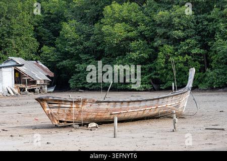 Krabi Thailand 19. Februar 2026. Ein verwittertes Holzboot liegt an einem Sandstrand in der Nähe eines kleinen Küstengebäudes. Stockfoto