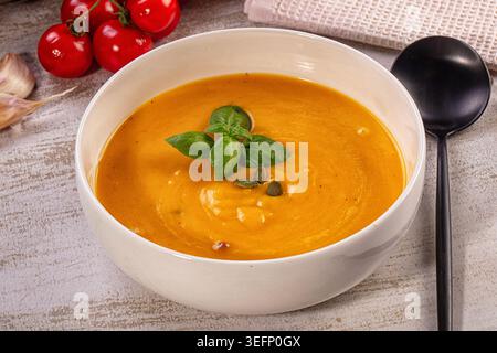 Nahaufnahme einer leuchtenden Orangencremesuppe in einer weißen Schüssel. Garniert mit frischen grünen Basilikumblättern, Kürbiskernen. Rustikaler Holzhintergrund mit rotem Chrom Stockfoto