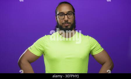 Mann mit neongrünem Hemd und Brille, Hände auf Hüften mit Unterarmen im Studio mit lila Hintergrund; Selbstvertrauen Kraft Fitness. Stockfoto