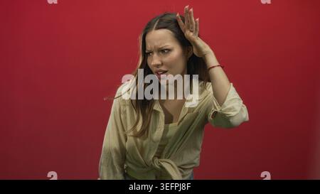 Frau mit Hand-zu-Ohr-Zuhören und Schielen im roten Studio mit gebundenem Hemd und Tanktop; Verwirrtheit Neugier. Stockfoto