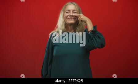Die Seniorin zeigt Finger an Nase und lacht in grünem Kleid vor rotem Studio-Hintergrund; verspielte Freude. Stockfoto