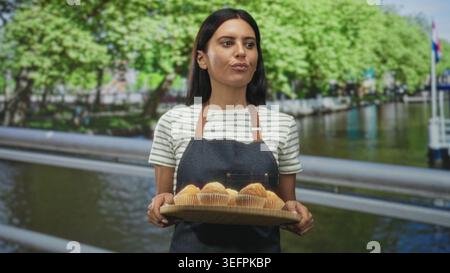 Bäckerin hält Tablett mit Muffins mit beiden Händen auf einer Straße am Kanal, präsentiert Gebäck; kuriose Gastfreundschaft. Stockfoto