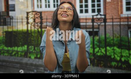 Frau zeigt mit erhobener Hand und gefaltetem Arm vor das Gebäude, trägt Brille und Jeansshirt; Verwirrung. Stockfoto