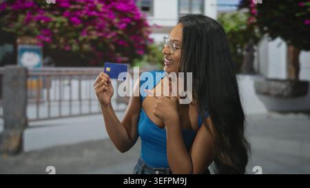 Junge Brünette, die eine Kreditkarte hält und auf der Straße mit dem Finger auf die Karte zeigt, die neben Geländer und Bougainvillea blau oben trägt; Selbstvertrauen Stockfoto